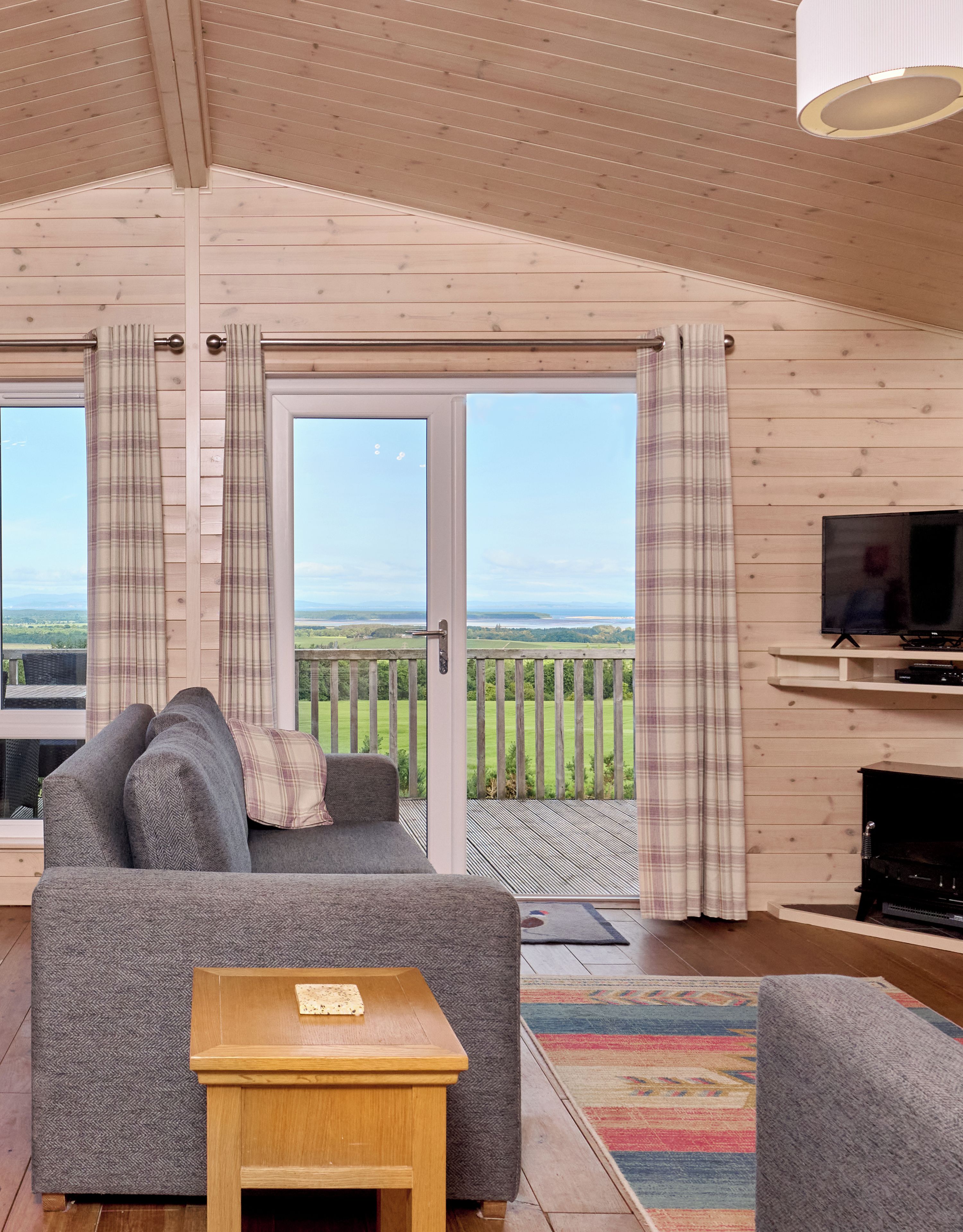 A cozy living room with a sofa, a small wooden table, and a television. There is a large sliding glass door leading to a balcony with a view of the countryside.