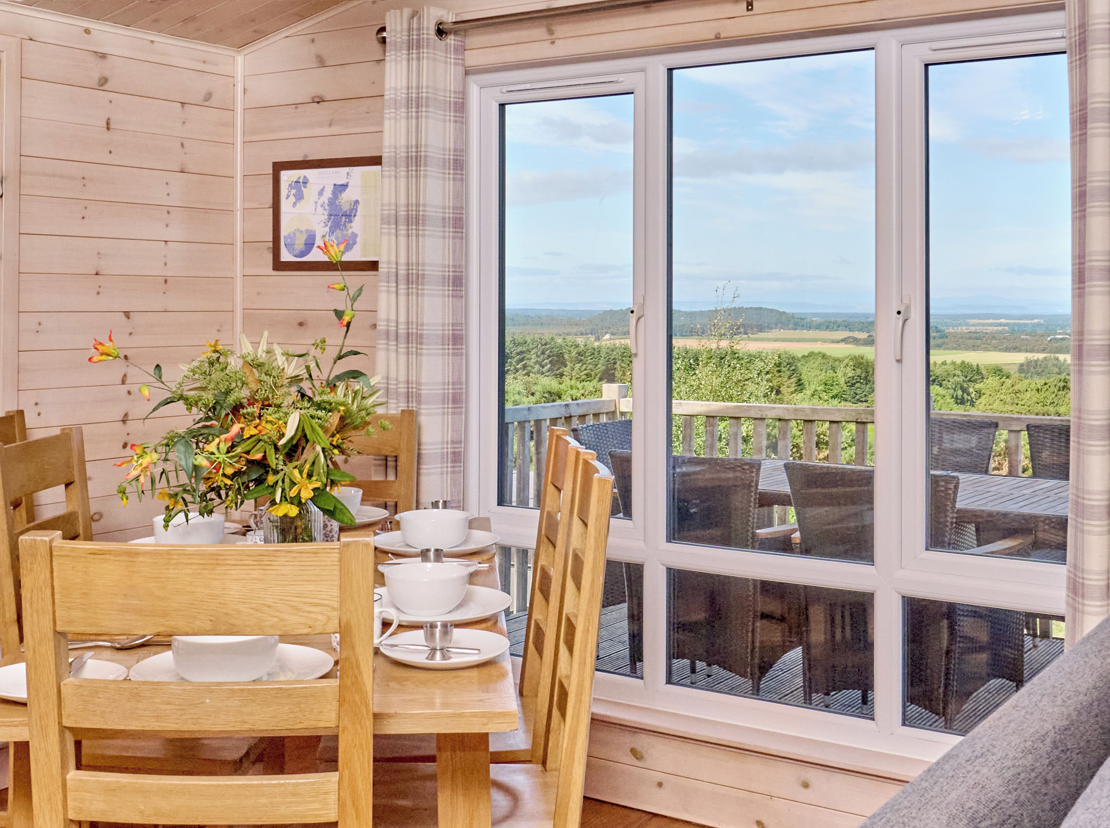 Un coin repas en bois, avec une table dressée pour deux personnes, un vase de fleurs et une vue sur un paysage verdoyant par de grandes fenêtres.