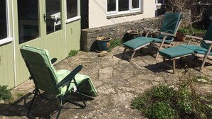 Une maison à deux étages avec un petit garage attenant. On y trouve deux chaises de jardin vertes et une table à l’extérieur.