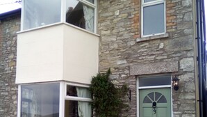 Une maison en pierre à deux étages, dotée d’une baie vitrée, d’une porte verte et d’un petit jardin agrémenté de plantes.