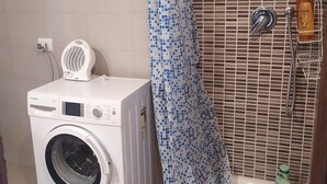 Kylpyhuoneessa on pyykinpesukone, sinivalkoruudullinen suihkuverho ja seinällä on hylly, jossa on hygieniatuotteita.