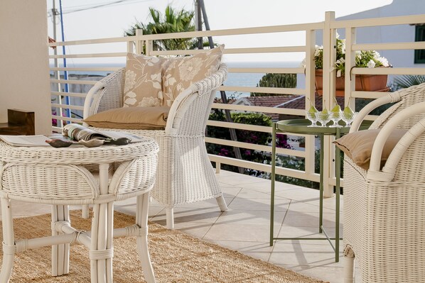Un coin salon aménagé avec des meubles en osier, comprenant une table et des chaises, une petite table avec des verres et un plateau, ainsi qu’une balustrade de balcon agrémentée de plantes en pot.