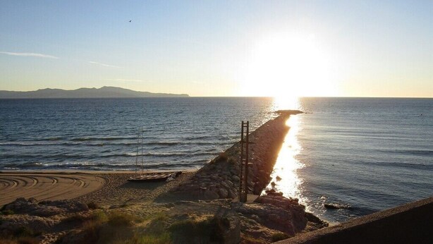 A coastal scene with a clear sky, a sun reflecting off the water, and a rocky shoreline.