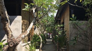 Un sendero estrecho bordeado de árboles y plantas en macetas, que conduce a un edificio con tejado de baldosas y pared de hormigón.