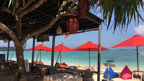 Un café frente a la playa con mesas al aire libre, sombrillas rojas y vistas al océano.