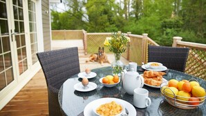Una mesa de comedor dispuesta con alimentos para el desayuno, un perro y vistas a una terraza y follaje verde.