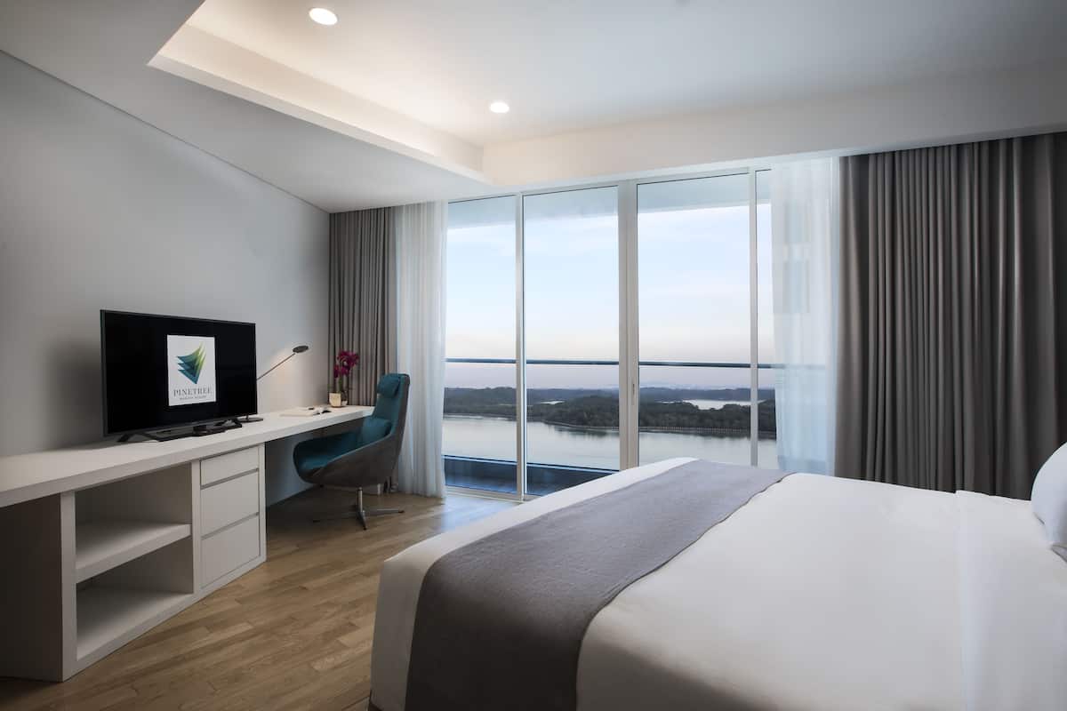 A modern hotel room with a large bed, a desk with a computer, a chair, and a large window offering a view of a body of water.