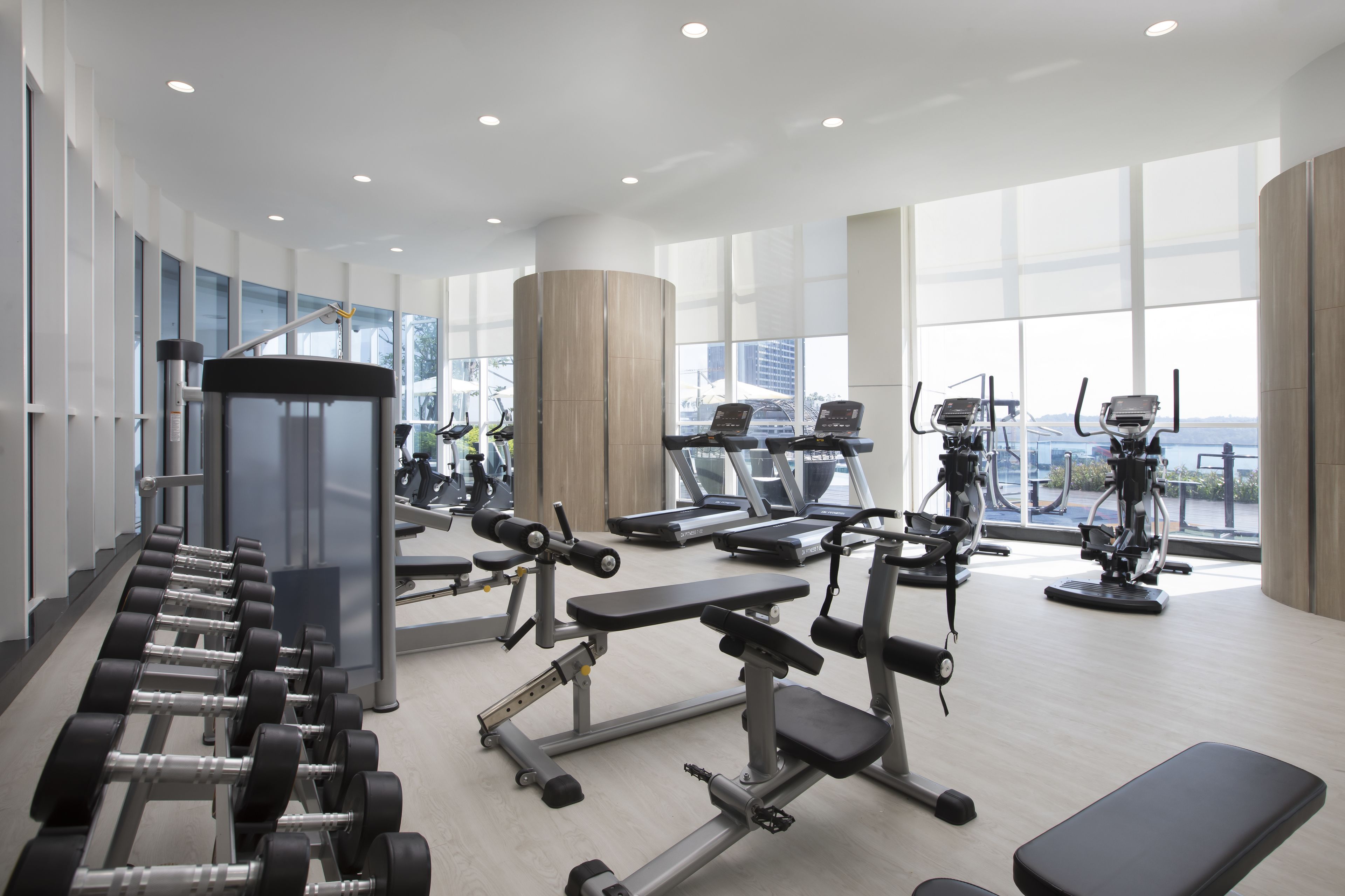 A modern gym with various exercise equipment, large windows, and a high ceiling.