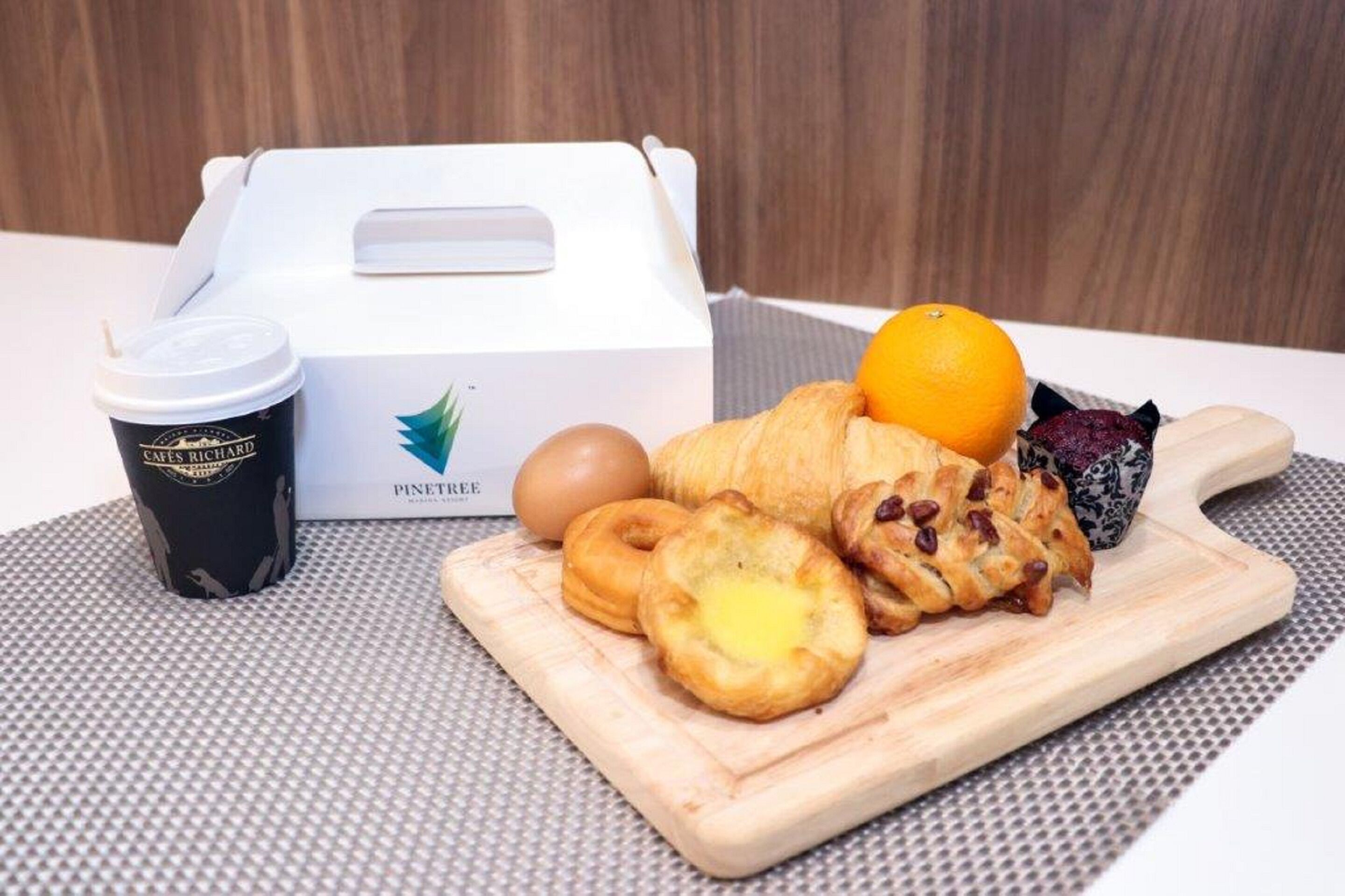 A breakfast setup with a box of pastries, a coffee cup, an orange, and a wooden cutting board.