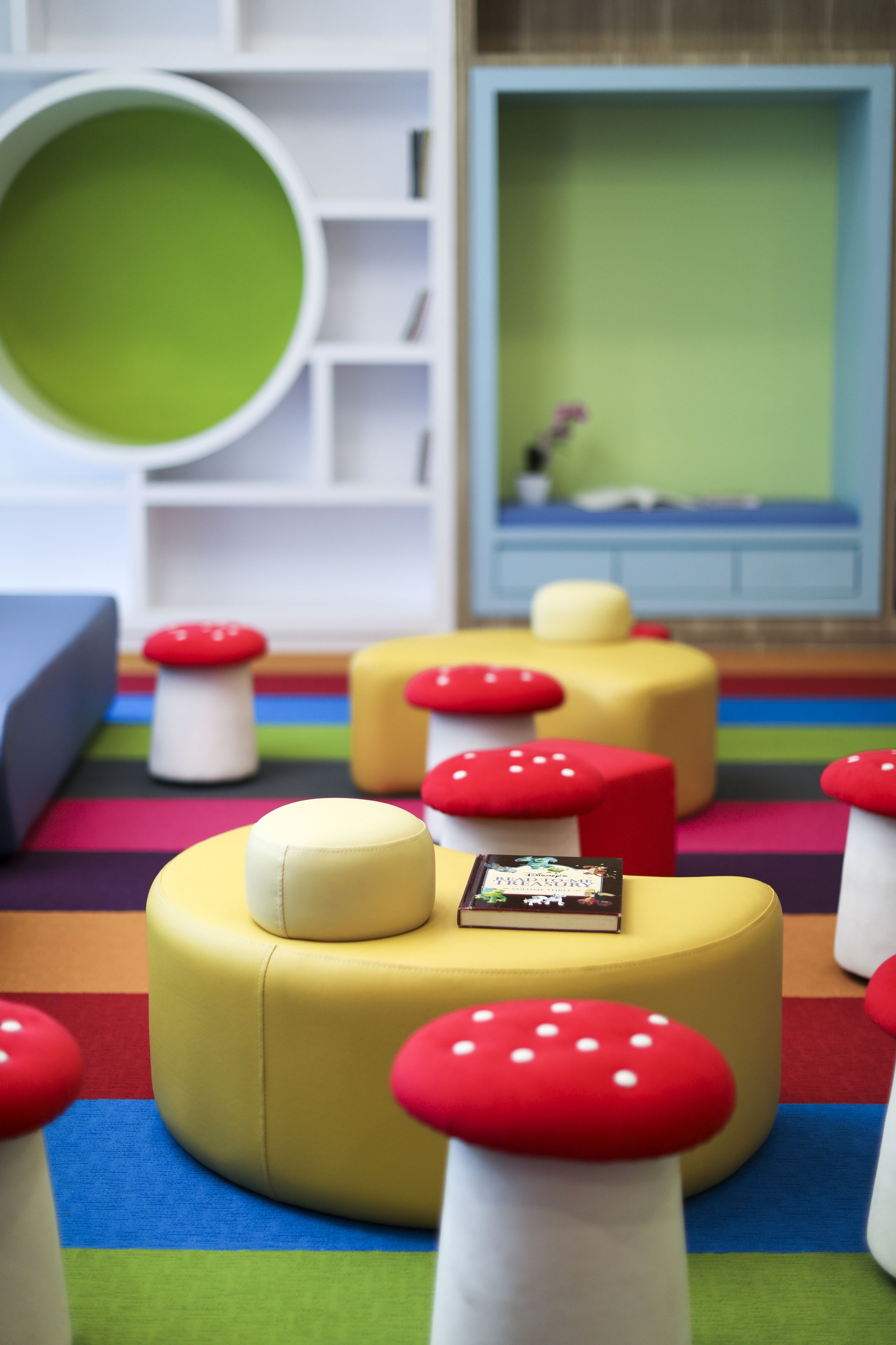A colorful playroom with round ottomans, some with books on them, and a green circular object on the wall.