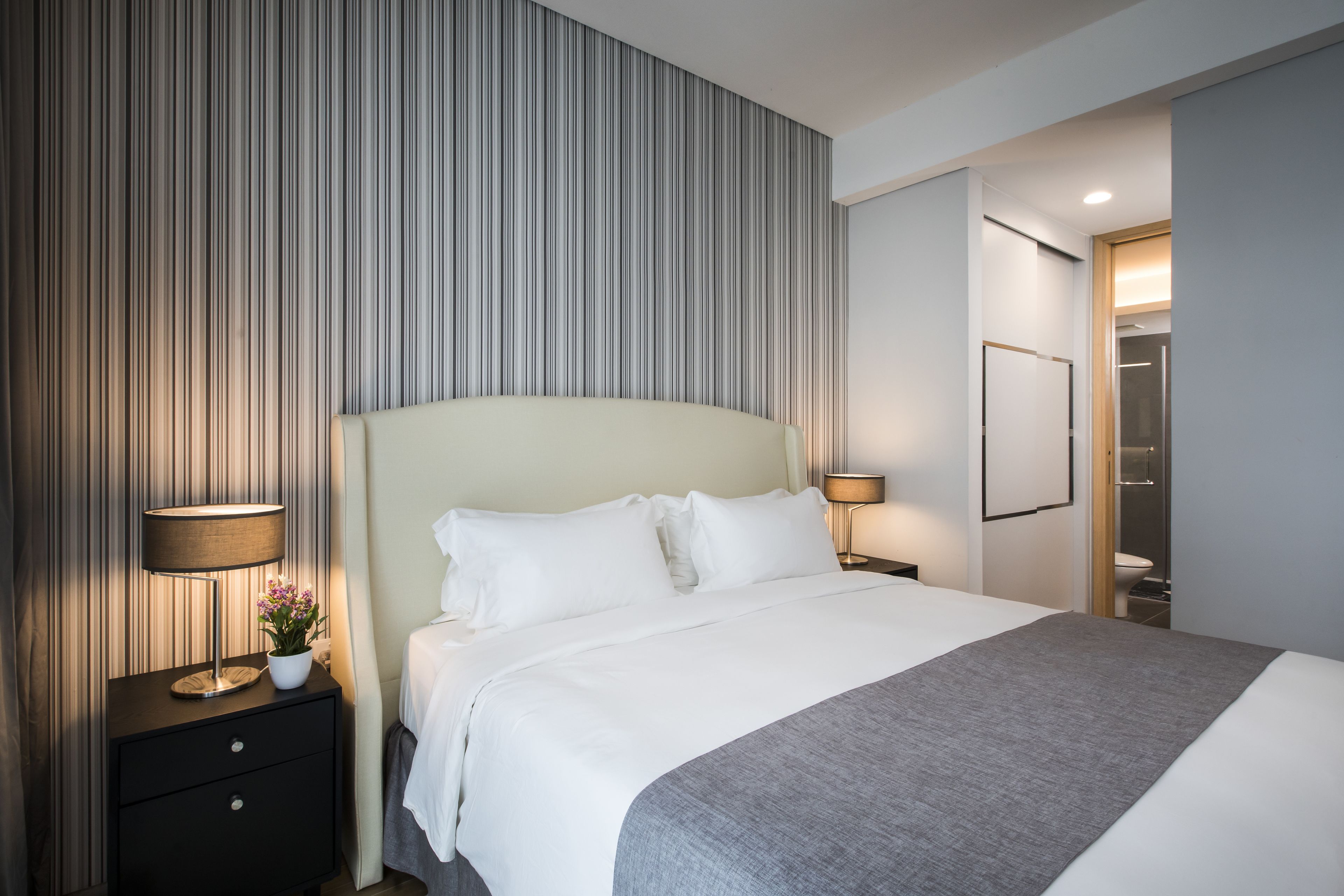 A hotel room with a large bed, two bedside lamps, a nightstand with a lamp and a flower, and a striped wall.