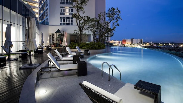 Un área de piscina en la azotea con sillones y sombrillas, rodeada de edificios modernos y un cielo despejado.