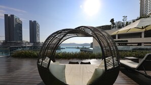 Silla de mimbre circular con cojines, una terraza de madera y vistas a edificios altos y un cuerpo de agua.
