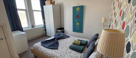 A bedroom with a bed, a wardrobe, a window with curtains, a wall with a floral pattern, and a lamp.