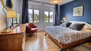 Ein Schlafzimmer mit einem hölzernen Bett, einem roten Sessel, einem Schreibtisch mit Lampe und einem großen Fenster mit Vorhängen.