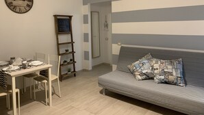 A room with a dining table set for four, a sofa with beach-themed pillows, a clock on the wall, and a shelf with plants.