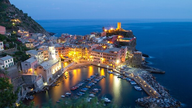 A coastal town at dusk with a marina, historic buildings, and a prominent tower.