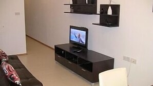 A living room with a dark brown entertainment unit, a flat-screen TV, a white chair, and a brown sofa with a patterned throw pillow. - Apartment in the center of Sliema (Tas-Sliema)