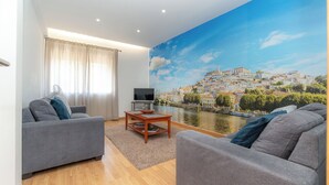 Una sala de estar moderna con un amplio mural panorámico de un río y un paisaje urbano, sofás grises, una mesa de centro de madera y un televisor de pantalla plana.