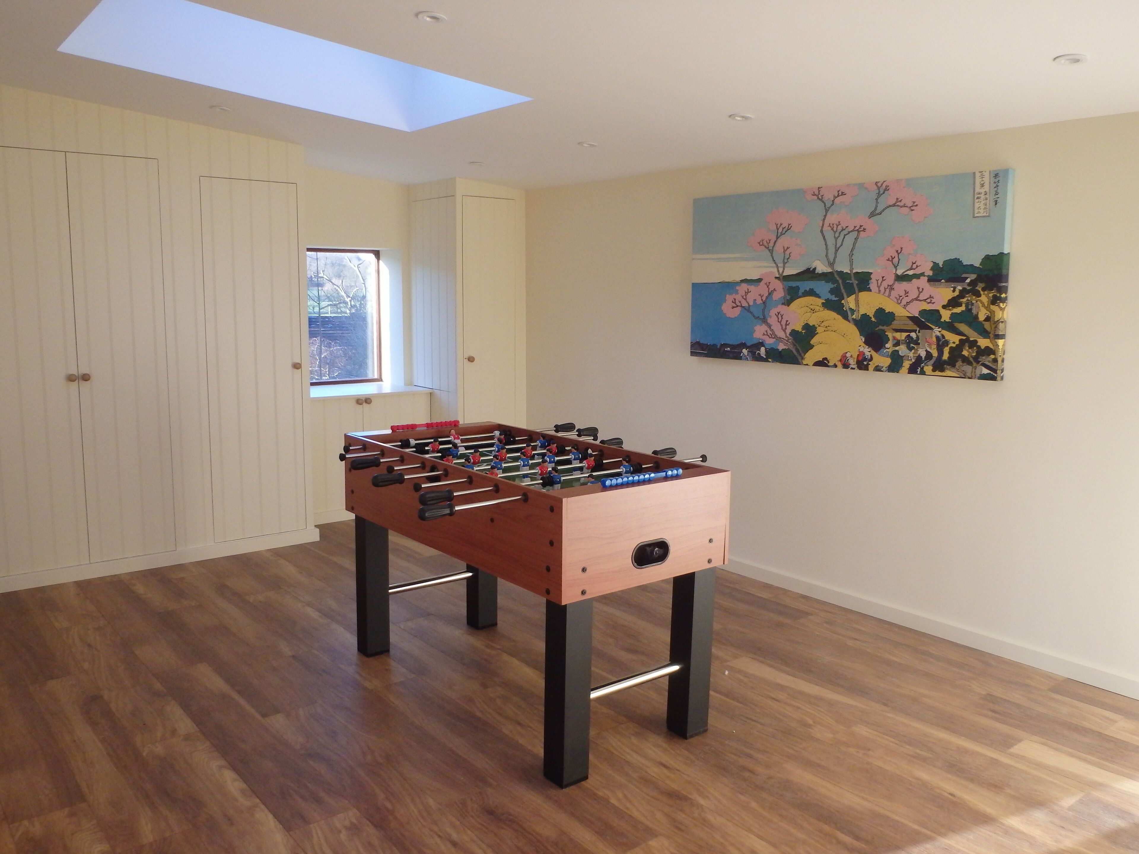 A room with a foosball table, wooden flooring, a skylight, a window, and a painting on the wall.