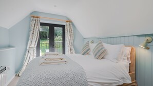 Une chambre avec un plafond en pente, un lit recouvert de draps blancs, des coussins à rayures et une vue sur un jardin, visible à travers une fenêtre ornée de rideaux.