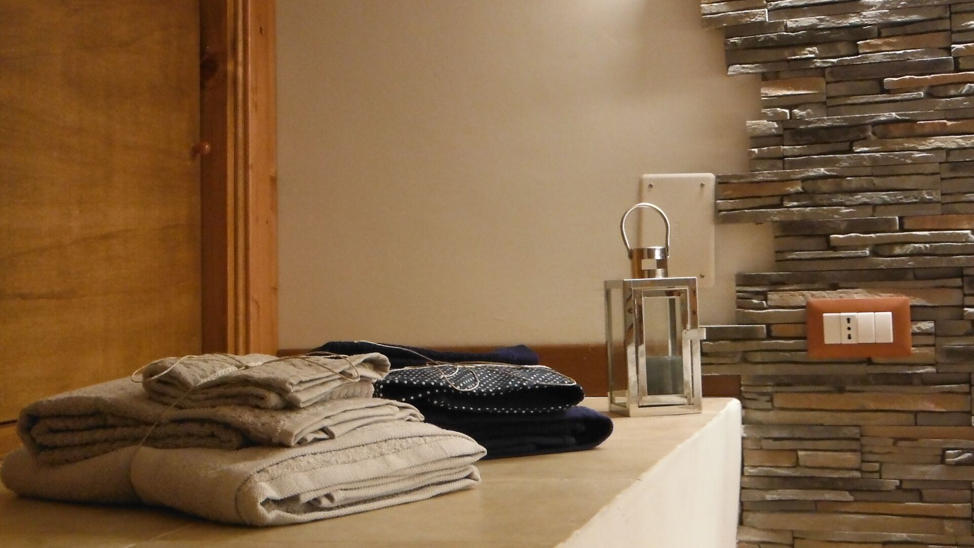 Un bagno con parete in pietra, un portascopevoli con asciugamani piegati, una piccola borsa nera e un corpo illuminante a parete.