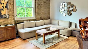 Uma sala de estar com piso de madeira, um sofá bege, uma mesa de centro de madeira, uma janela grande com vista para vegetação e uma parede de tijolos.