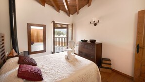 A bedroom with a wooden ceiling, a bed with a white quilt and two pillows, a dresser, a chair, and a view of the outdoors through an open door.