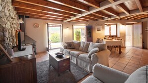 A cozy living room with a stone wall, wooden beams, a sofa, a coffee table, and a dining area with a table and chairs.