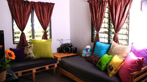 A cozy room with a wooden bench and a sofa, both with colorful cushions. There is a small table with a radio and a potted plant. - The Retreat Homestay (Tanjung Malim)