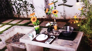 An outdoor dining table set with a bottle, glasses, and cutlery, surrounded by plants and flowers. - The Retreat Homestay (Tanjung Malim)