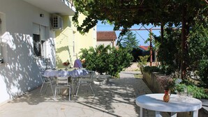 An outdoor patio with a round table and chairs, a small round table with a vase and wine glasses, and a garden area with a tree and shrubs.