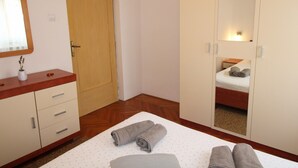 A bedroom with a wooden door, a mirror, a bed with a mattress and pillows, and a bedside table with a lamp.