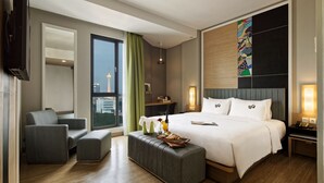 Una habitación de hotel moderna con una cama grande, un área de descanso con un sofá y sillón, una mesita con una bandeja de desayuno, y vista a la ciudad desde la ventana.