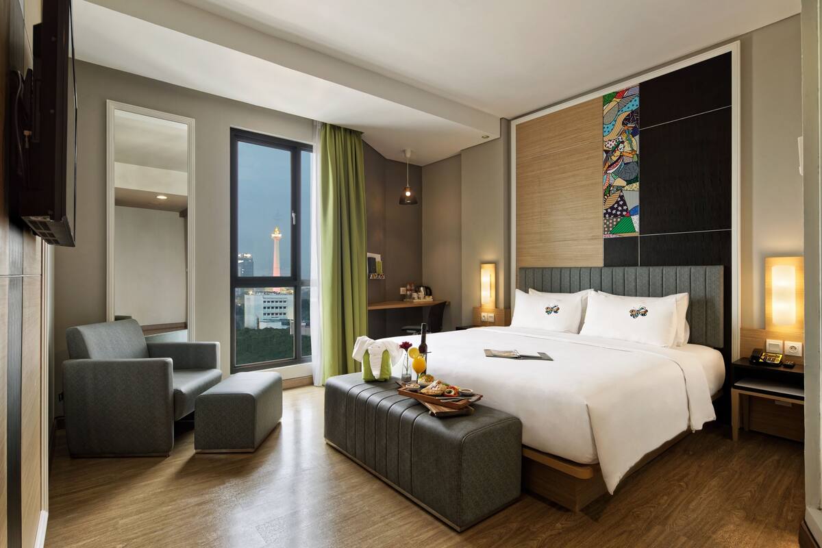 A modern hotel room with a large bed, a seating area with a sofa and armchair, a small table with a tray of breakfast, and a city view through the window.
