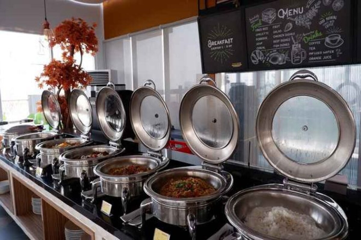 Un área de bufé con diversas opciones de comida en recipientes de acero inoxidable, un panel con el menú arriba y una planta decorativa a la izquierda.