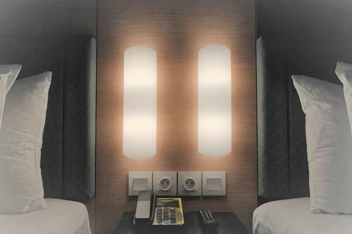 Una habitación de hotel con cama, dos mesitas de noche y luces fijadas a la pared.