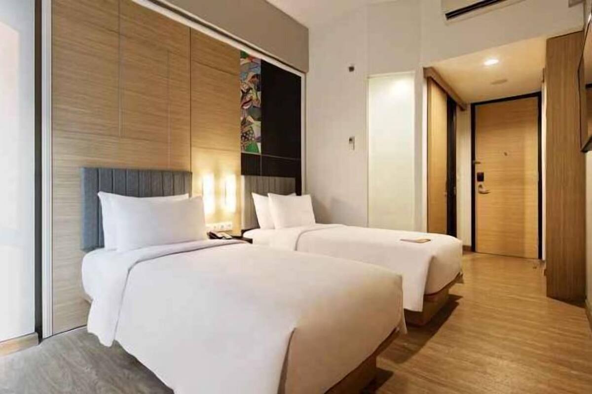 A hotel room with two beds, a wooden wall panel, a colorful artwork, and a door leading to another room.