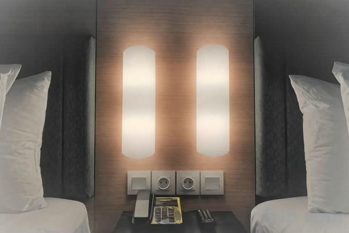 A hotel room with two beds, a nightstand with a telephone and remote controls, and wall-mounted lighting.