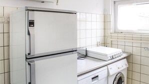 A compact kitchen with a refrigerator, a washing machine, and a window with blinds.