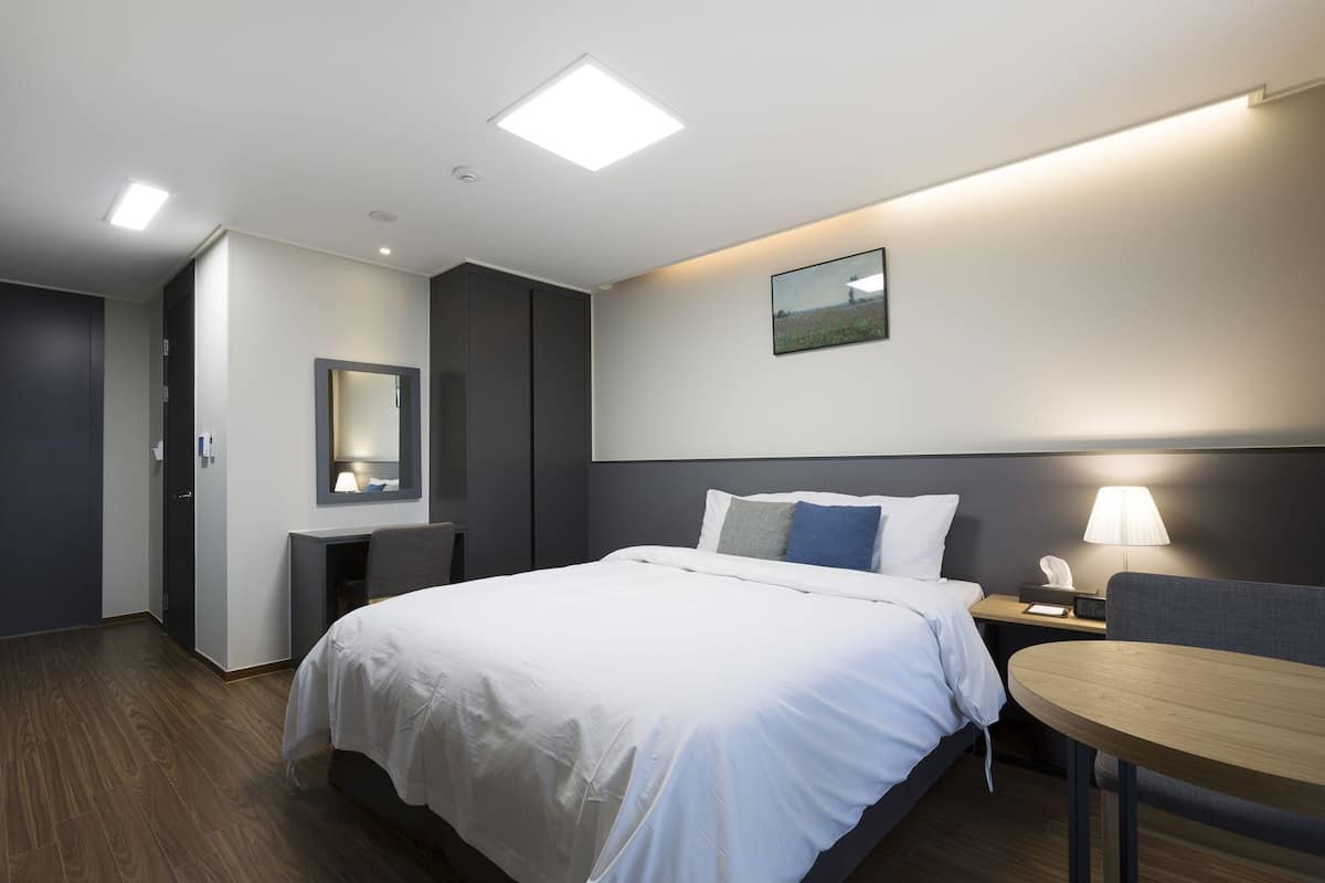A hotel room with a bed, bedside table, lamp, desk, chair, mirror, and a picture on the wall.