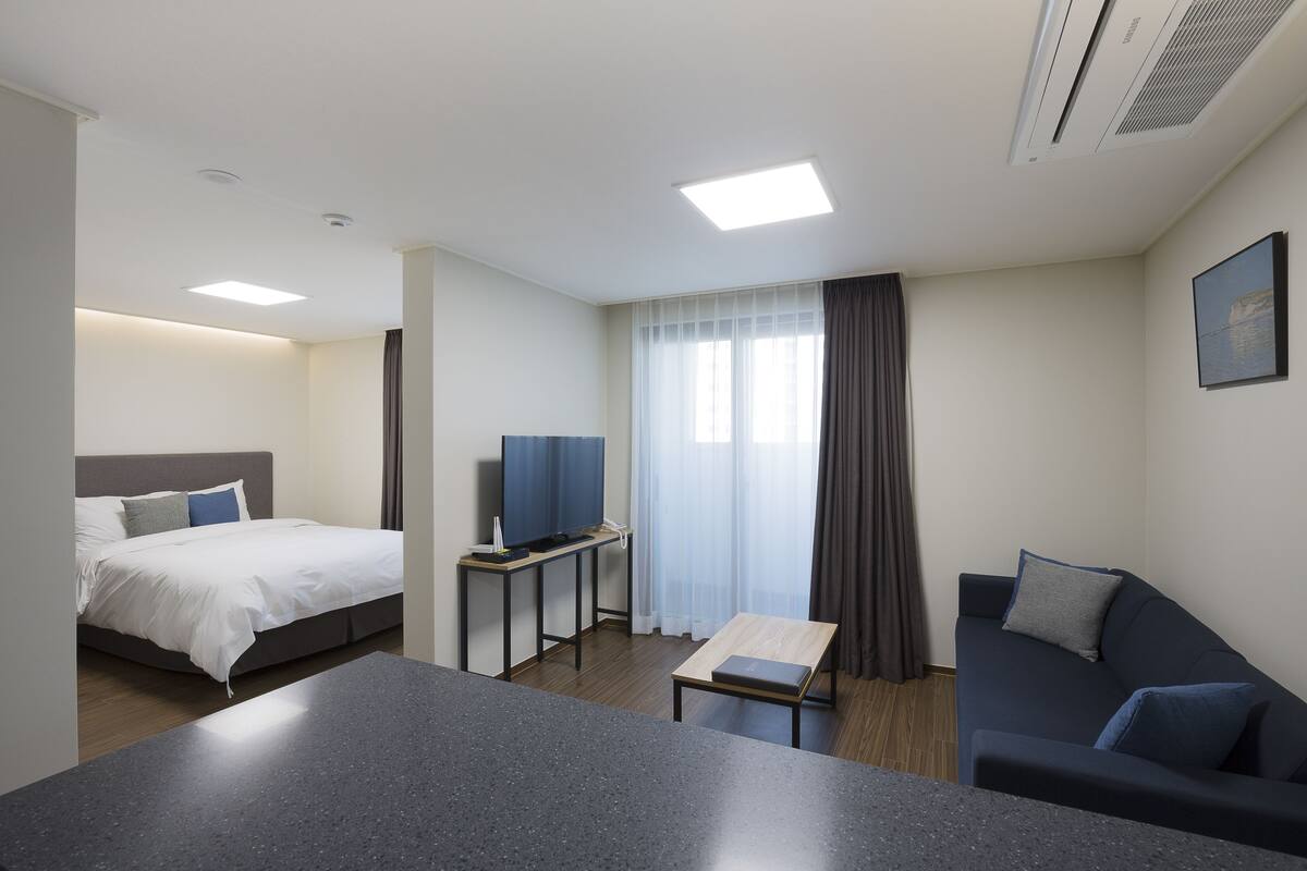 A modern hotel room with a bed, a sofa, a small table, a TV, and a window with curtains.
