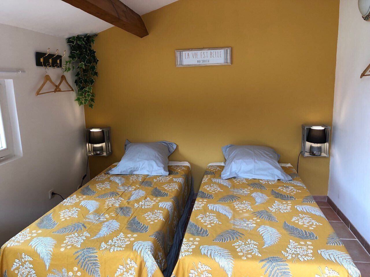 A room with two beds, each with a yellow floral bedspread and matching pillows. There are wall-mounted lamps on either side of the beds, and a sign above the beds that reads "LA VIE EST BELLE AU SOLEIL".
