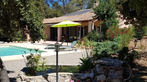 A pool area with a covered patio, a stone planter with flowers, and a house with a tiled roof.