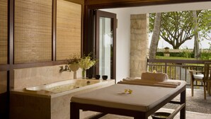 Un cuarto de spa con una bañera grande, una camilla de masaje y vistas a un jardín y un árbol afuera.