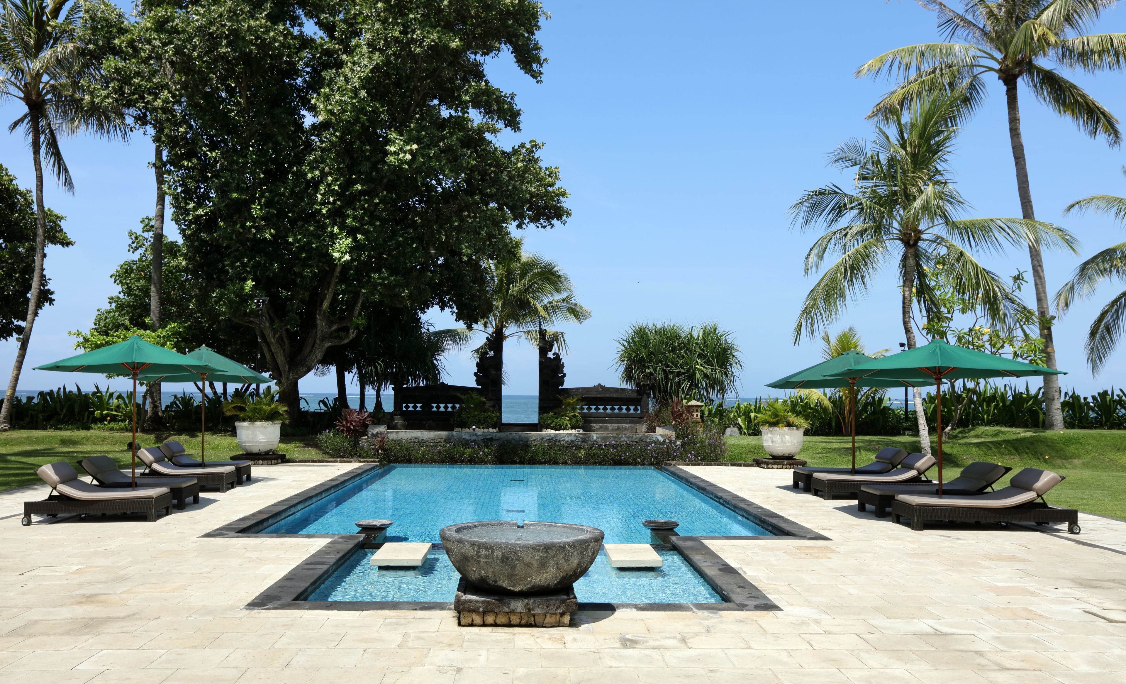 A pool area with lounge chairs and umbrellas, surrounded by palm trees and a view of the ocean.