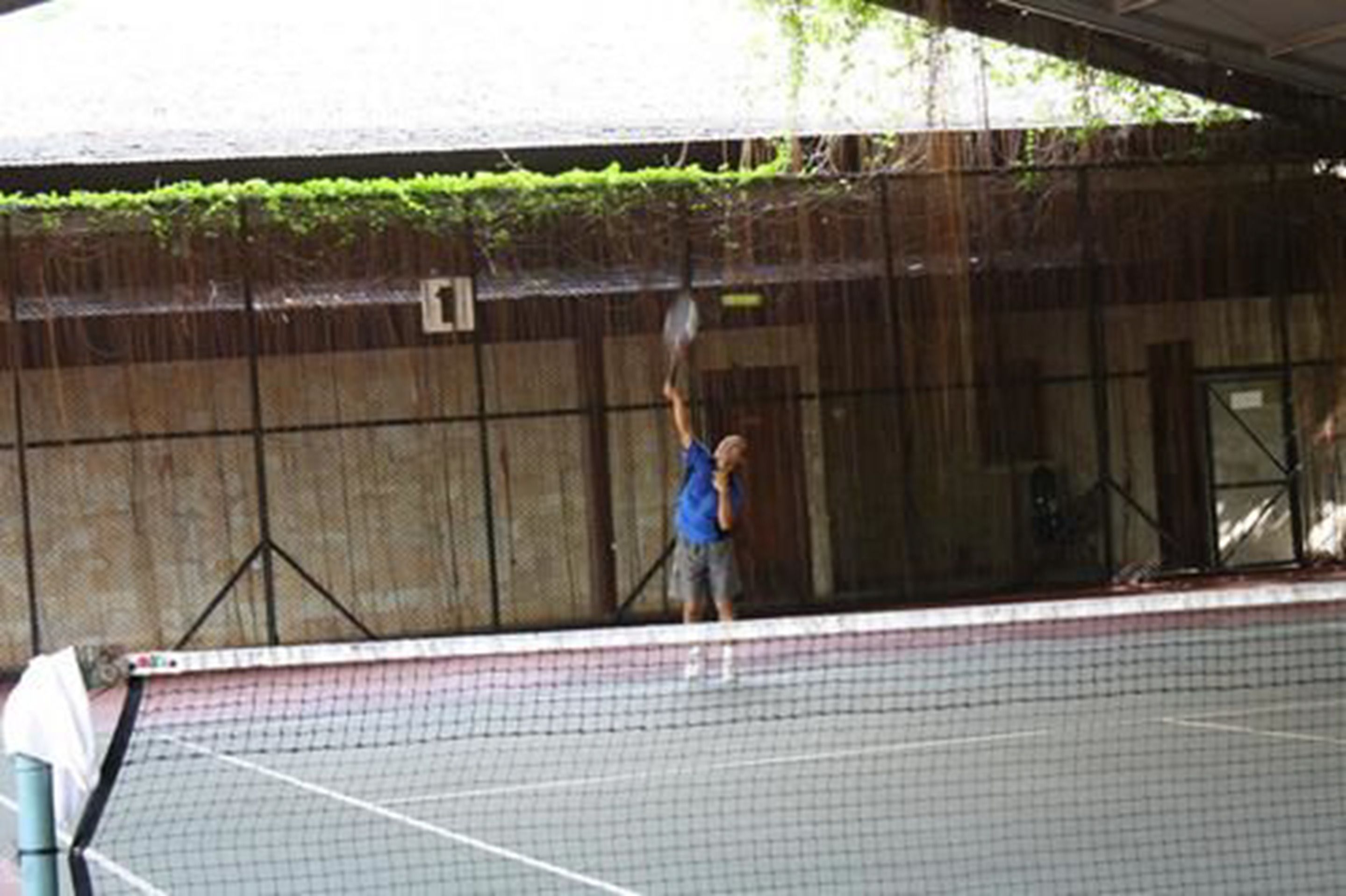 Una cancha de tenis con una red, un jugador con camisa azul y un área techada.