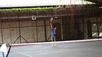 Una cancha de tenis con una red, un jugador con camisa azul y un área techada.