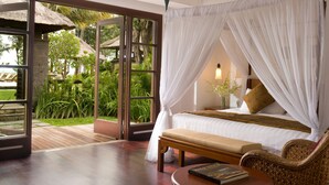 Un dormitorio con una cama grande, un banco, una silla de mimbre y vistas a un jardín a través de un ventanal.