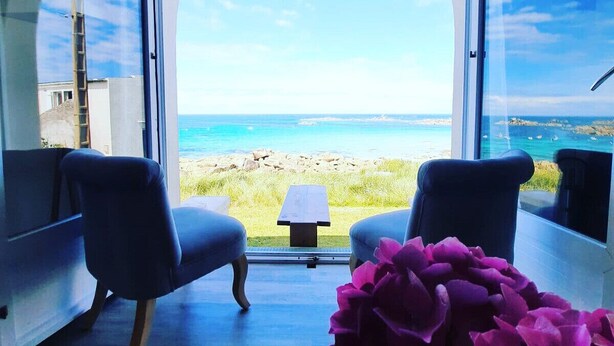 Ein Zimmer mit einem großen Bogenfenster mit Blick auf Strand und Meer. Zwei Stühle und ein kleiner Tisch stehen im Raum.